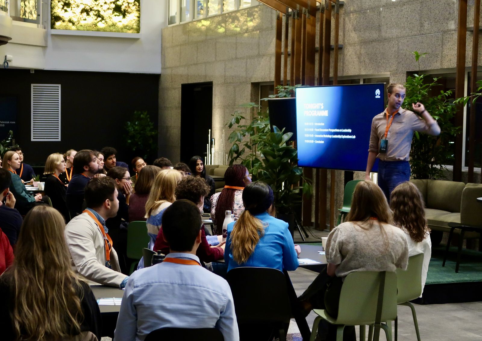 A speaker presenting the evening's programme to a full, seated audience in a plant-filled atrium at The VALE Symposium